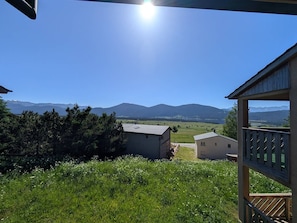 Uitzicht vanuit een overdekt buitengebied met een houten terras, op een grasveld en verre bergen onder een heldere lucht.