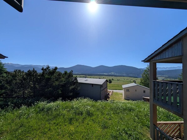 Vue depuis un espace extérieur abrité doté d’une terrasse en bois, donnant sur un champ verdoyant et des montagnes lointaines, le tout sous un ciel dégagé.