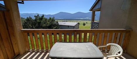 Una terraza de madera con una mesa y sillas, vista a montañas y un edificio al fondo.