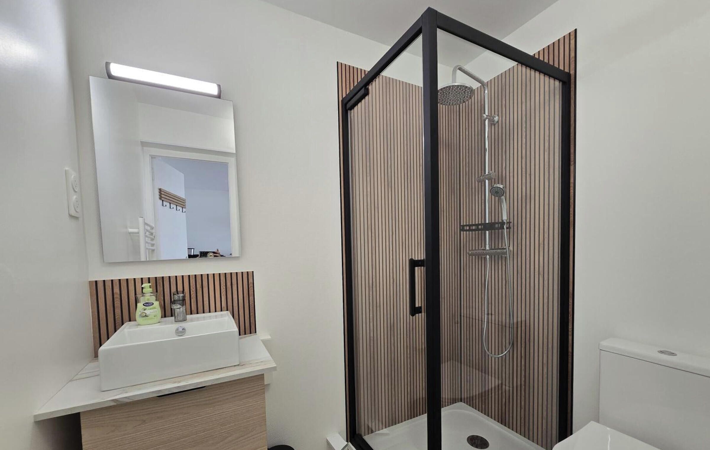 A modern bathroom with a glass-enclosed shower, a white sink with a wooden vanity, and a mirror with a light above it.