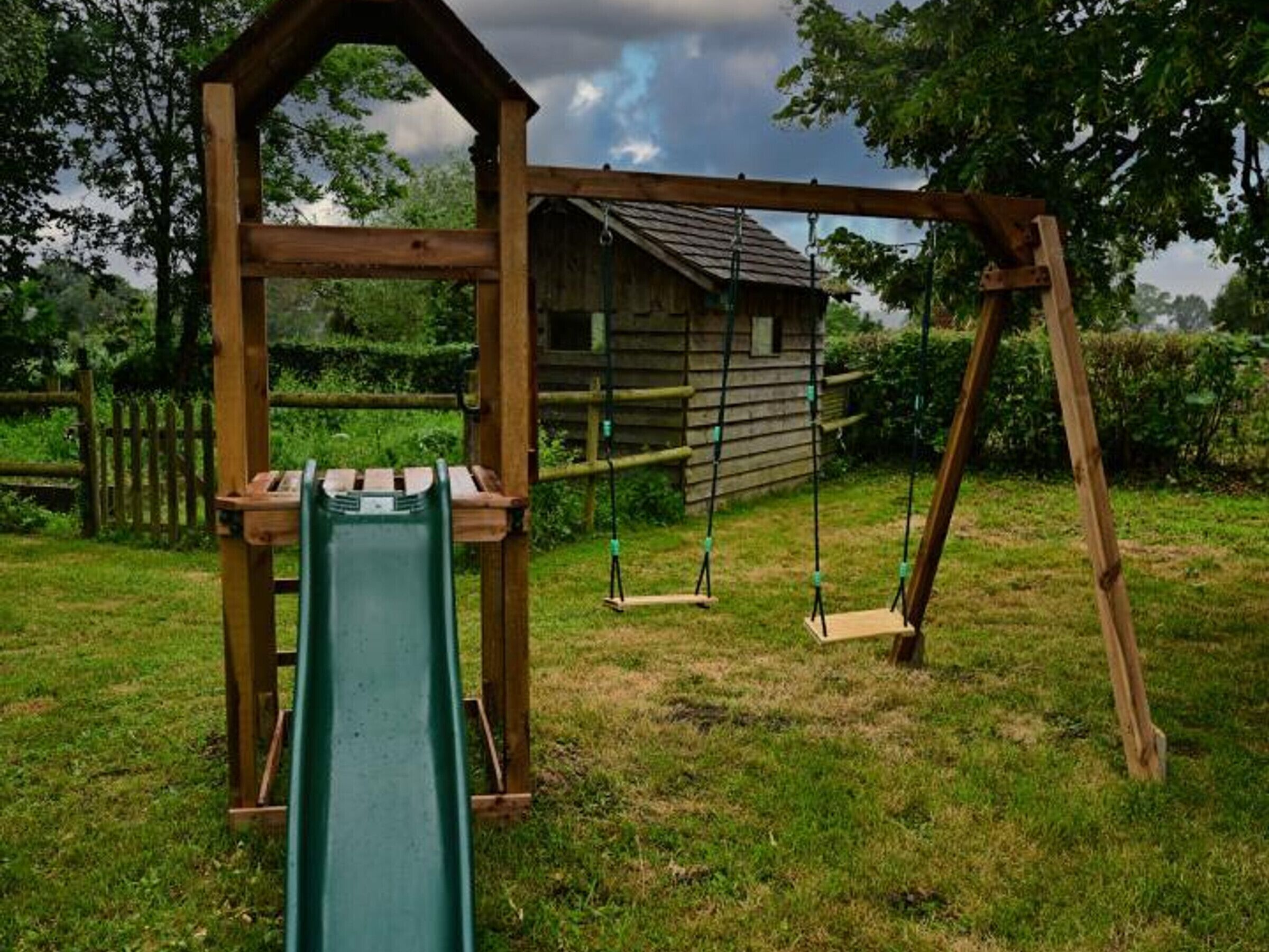 A wooden swing set with a slide, a small wooden structure, and a fenced area.