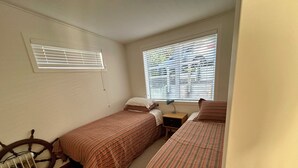 A small, single-room accommodation with two beds, a radiator, a window with blinds, and a wooden headboard.