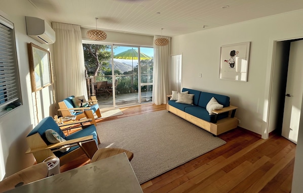 A spacious living room with a blue sofa, wooden furniture, a large window with curtains, and a view of the outdoors. - Cosy cabin style bach, immersed in native bush, in beautiful Banks Peninsula (Little Akaloa)