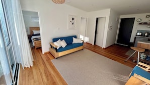 A modern living room with a blue sofa, wooden flooring, a hanging light fixture, and a kitchen area with a kettle visible.