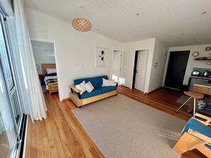Una sala de estar moderna con un sofá azul, suelo de madera, una lámpara colgante y una zona de cocina con una tetera a la vista.