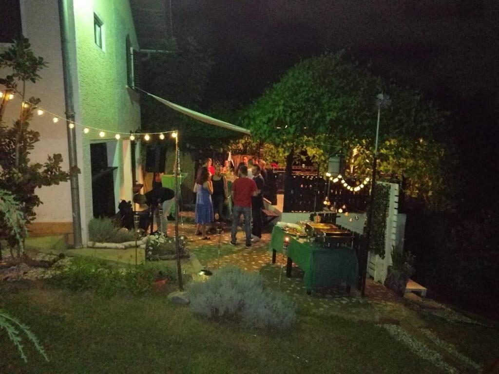 Un événement en plein air où des personnes sont réunies autour de tables sous des guirlandes lumineuses, un bâtiment avec une porte verte et un espace verdoyant avec des plantes.