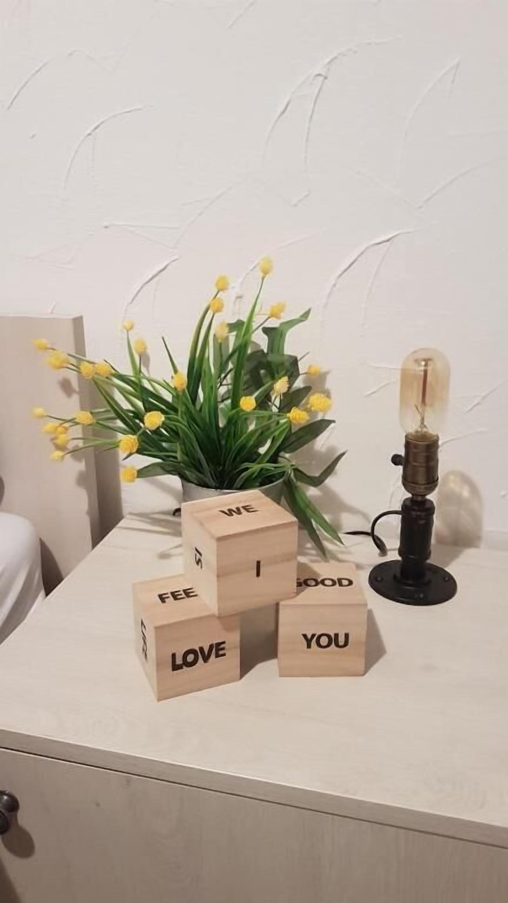 Uma mesa de cabeceira com uma planta em vaso, um abajur e blocos de madeira formando a frase 'WE LOVE YOU'.