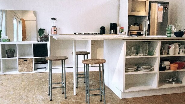 Una cocina con barra, taburetes, estantes con vajilla y una licuadora.