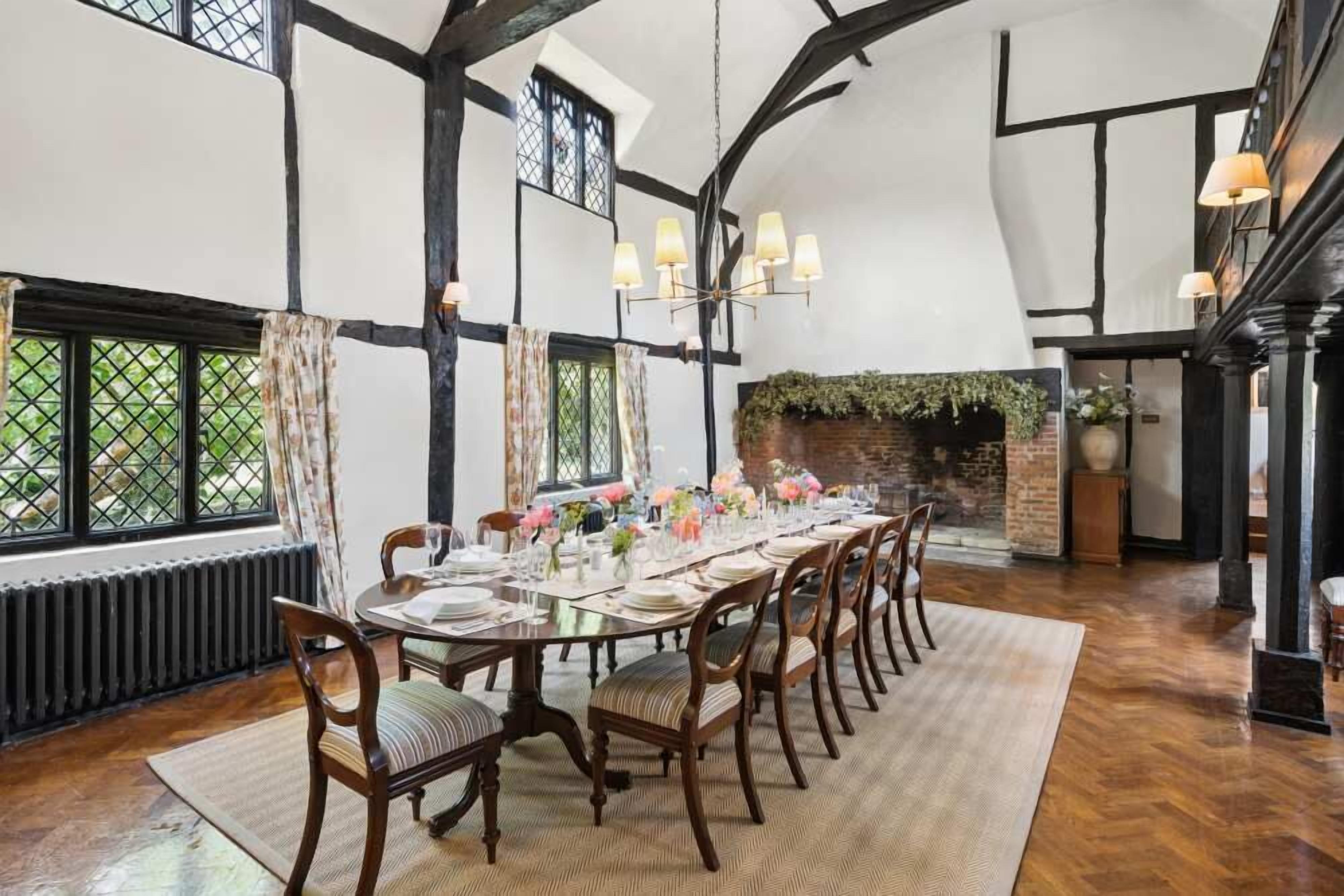 A dining room with a long table set for six, wooden chairs, a chandelier, and a brick fireplace.