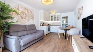 Un soggiorno moderno con un divano grigio, un tavolo da pranzo con sedie, una zona cucina e una TV sulla destra.