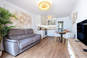 Ein modernes Wohnzimmer mit einem grauen Sofa, einem Esstisch mit Stühlen, einer Küchenzeile und einem Fernseher rechts.