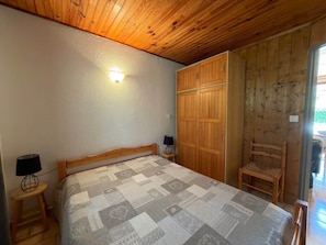 A bedroom with a wooden ceiling, a bed with a patterned bedspread, a wardrobe, a chair, and a nightstand with a lamp. - In the heart of the Verdon, with a terrace and garden! (Castellane)
