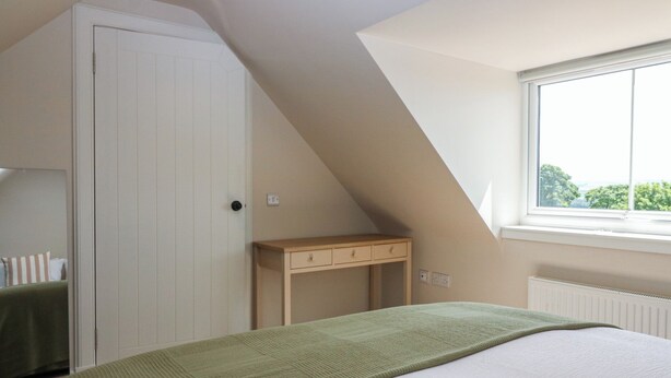 A bedroom with a sloped ceiling, a bed with a green blanket, a wooden nightstand, and a window with a view.