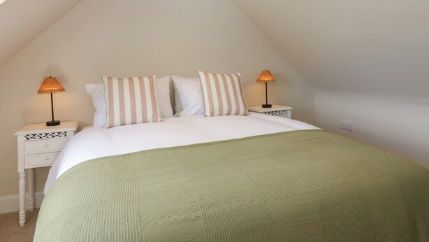 A bedroom with a sloped ceiling, a bed with striped pillows, two bedside lamps, and a green throw blanket. - Shepherds Cottage (Haddington)
