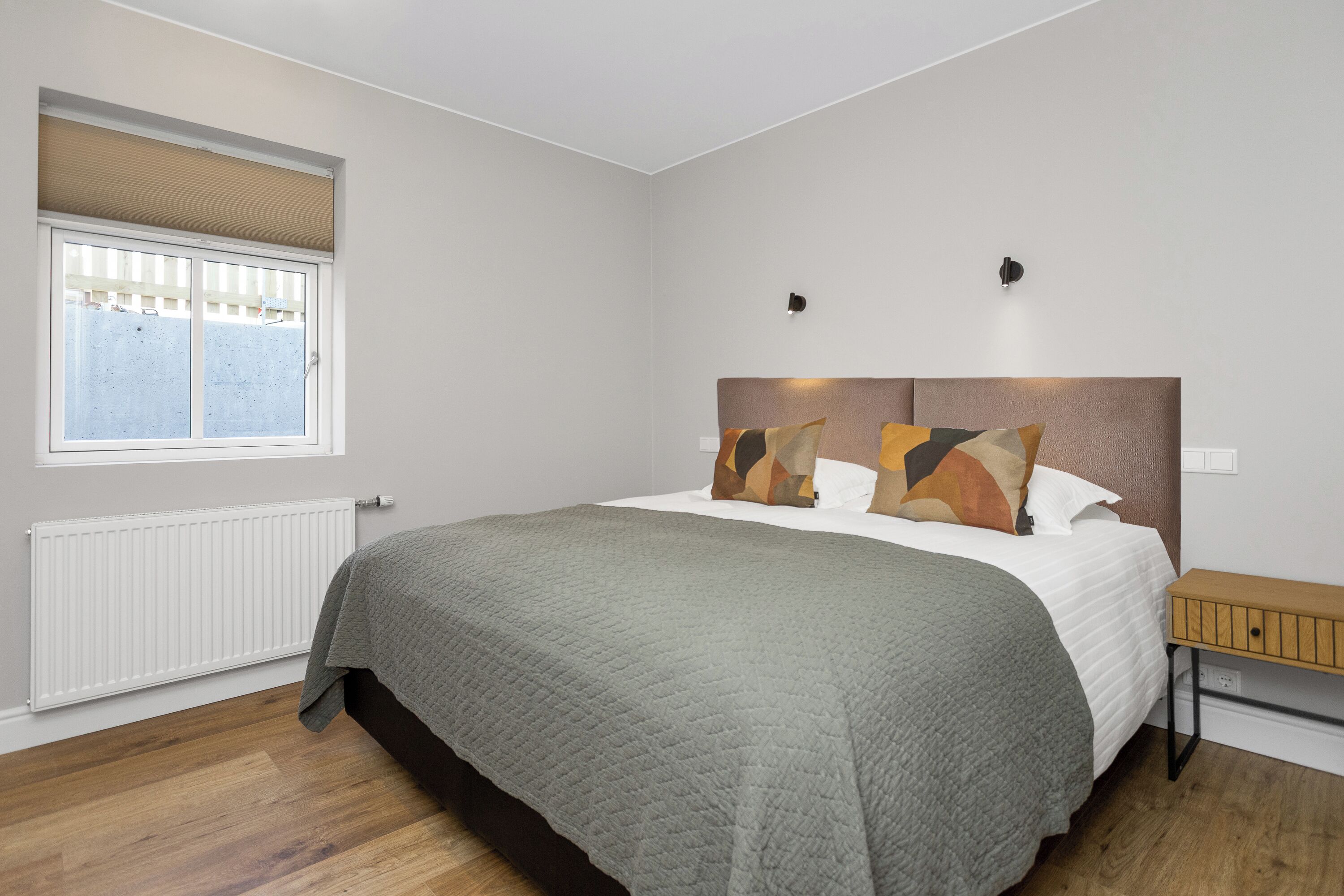 Ein Schlafzimmer mit Bett, einem Fenster mit Jalousien, einem Nachttisch und einer Wandlampe.