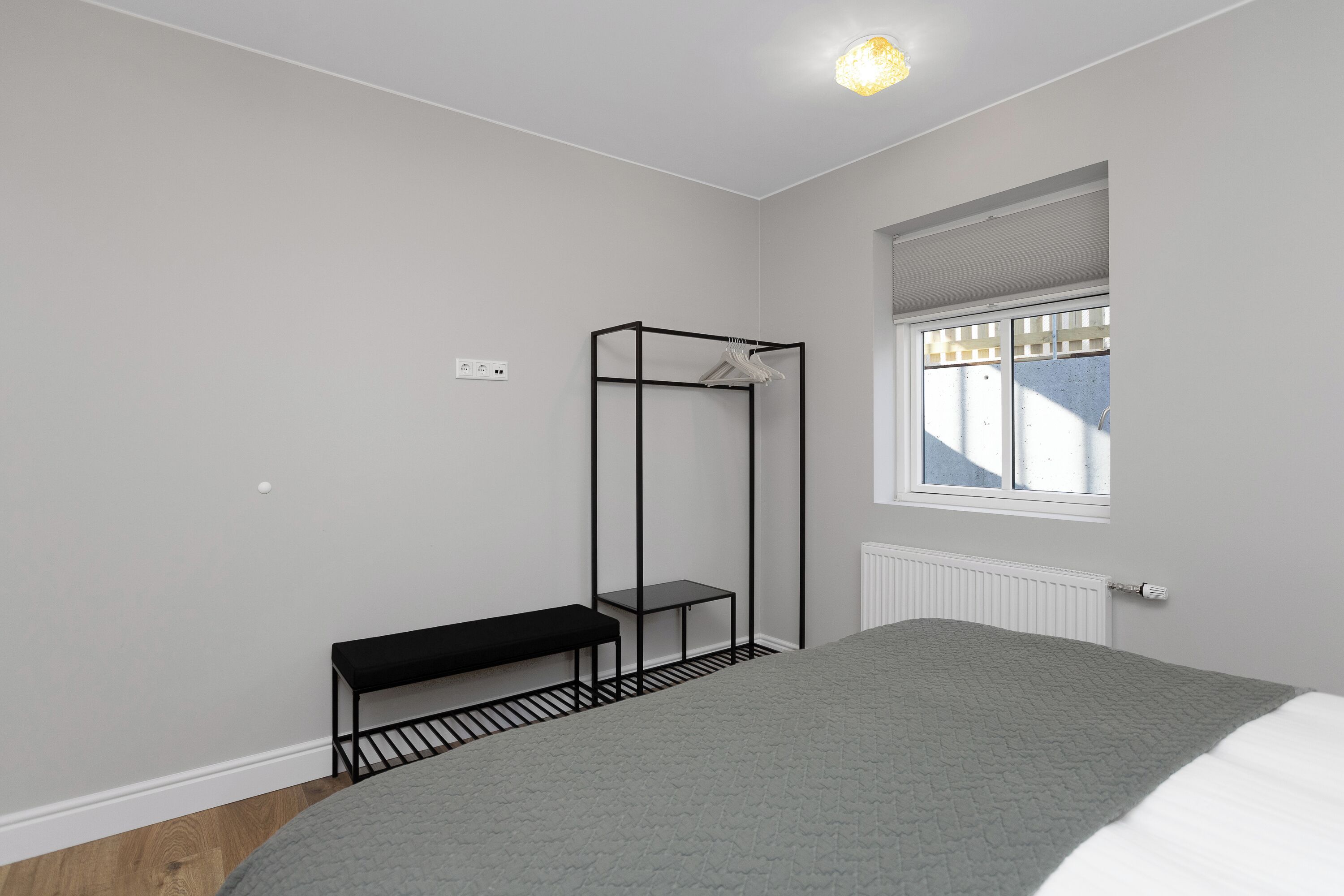 A bedroom with a bed, a bench, a metal shelf, a radiator, and a window.