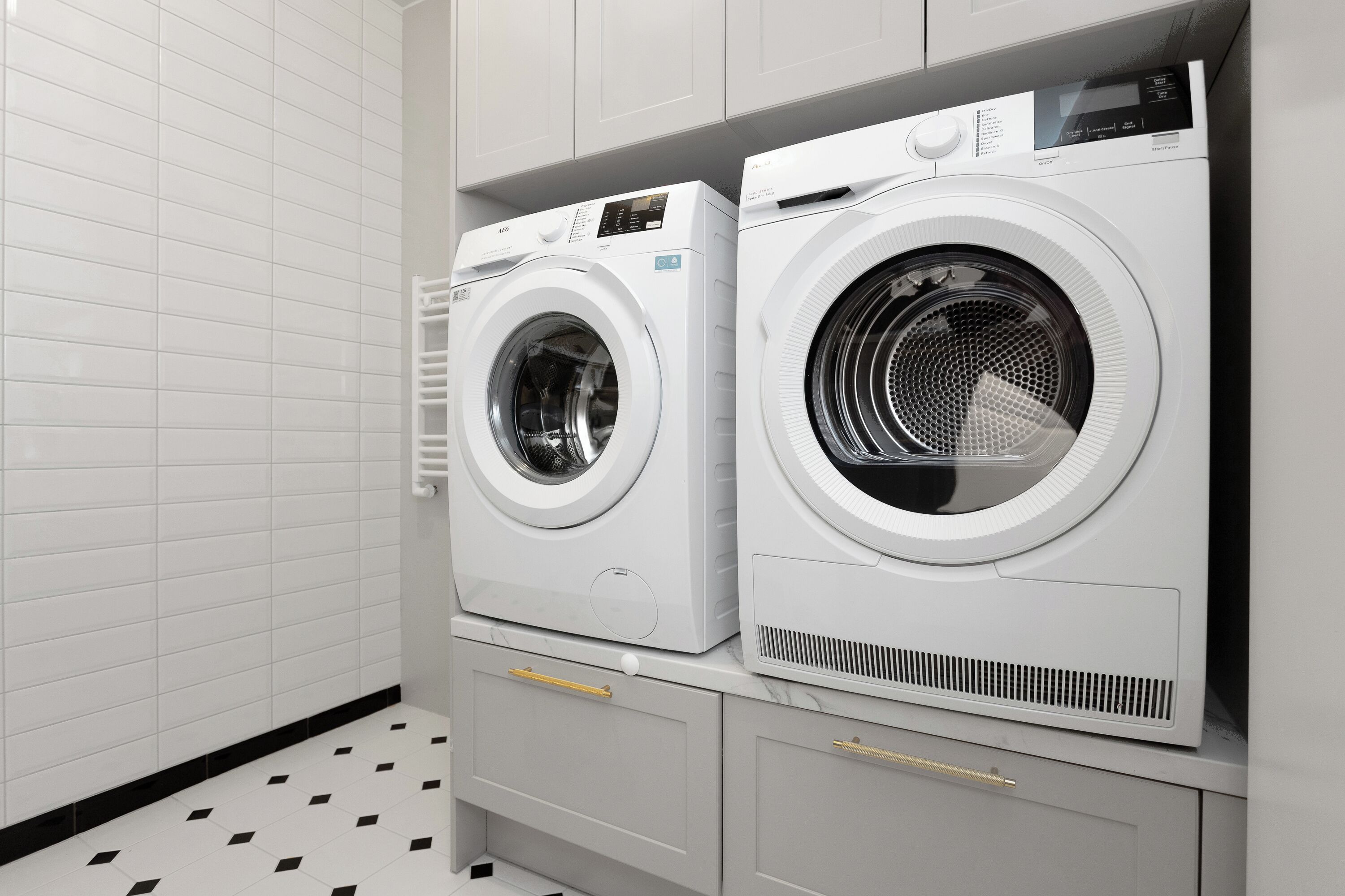 A laundry room with two front-loading washing machines, a tiled floor, and grey cabinets with drawers.