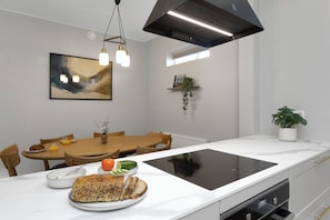 Eine moderne Küche mit weißer Arbeitsplatte, schwarischem Induktionskochfeld, einem Holztisch und Pendelleuchten.