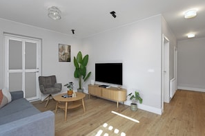A modern living room with a grey sofa, a wooden coffee table, a grey armchair, a large green plant, a flat-screen TV, and a wooden cabinet. - Aska Selfoss (Selfoss)