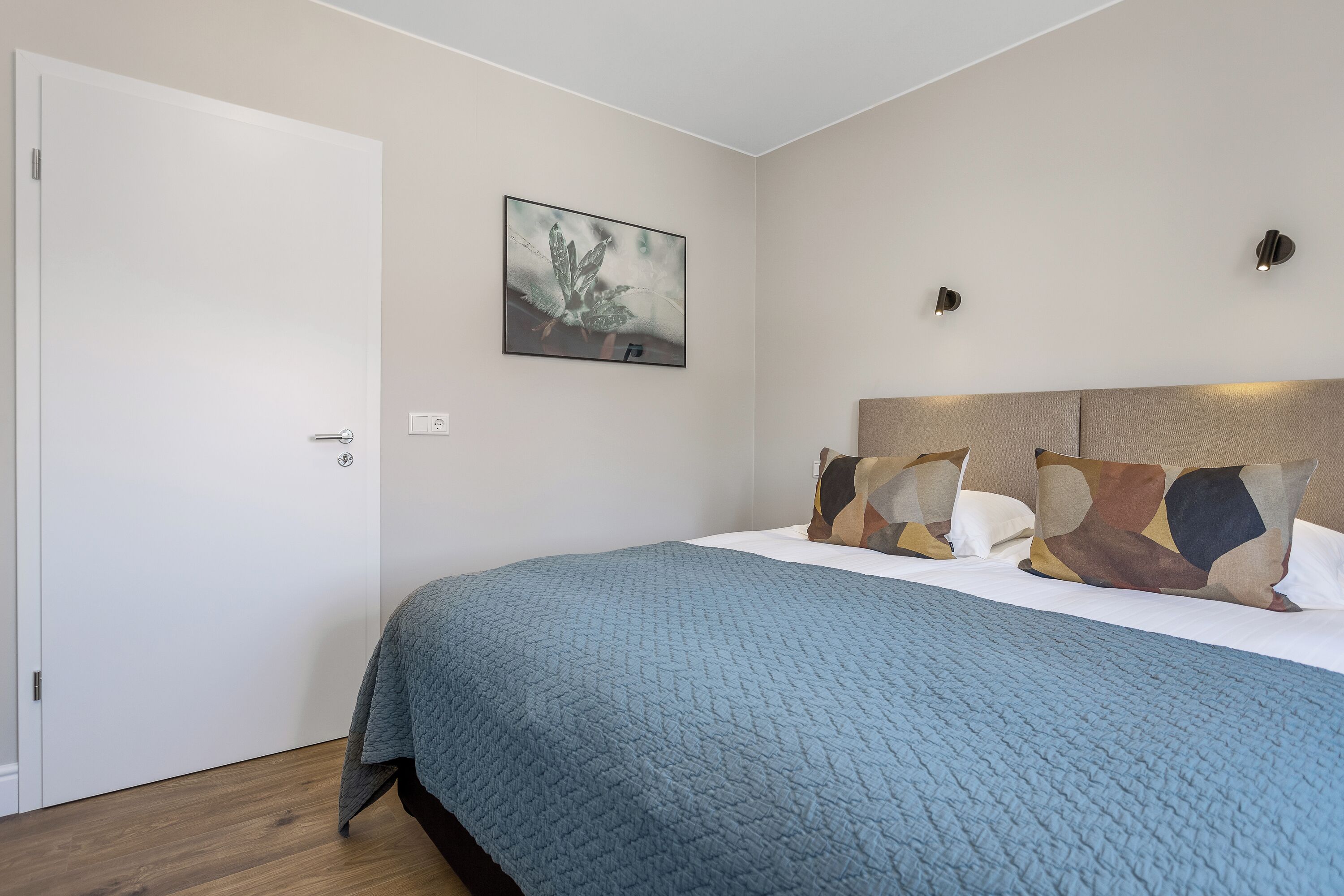 A bedroom with a bed, a door, a wall-mounted light, and a framed picture.
