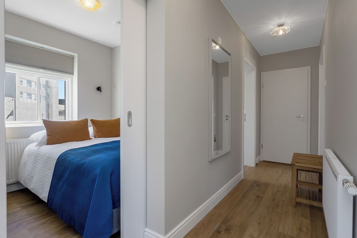 Um quarto com cama, janela, espelho e corredor que leva a uma porta.