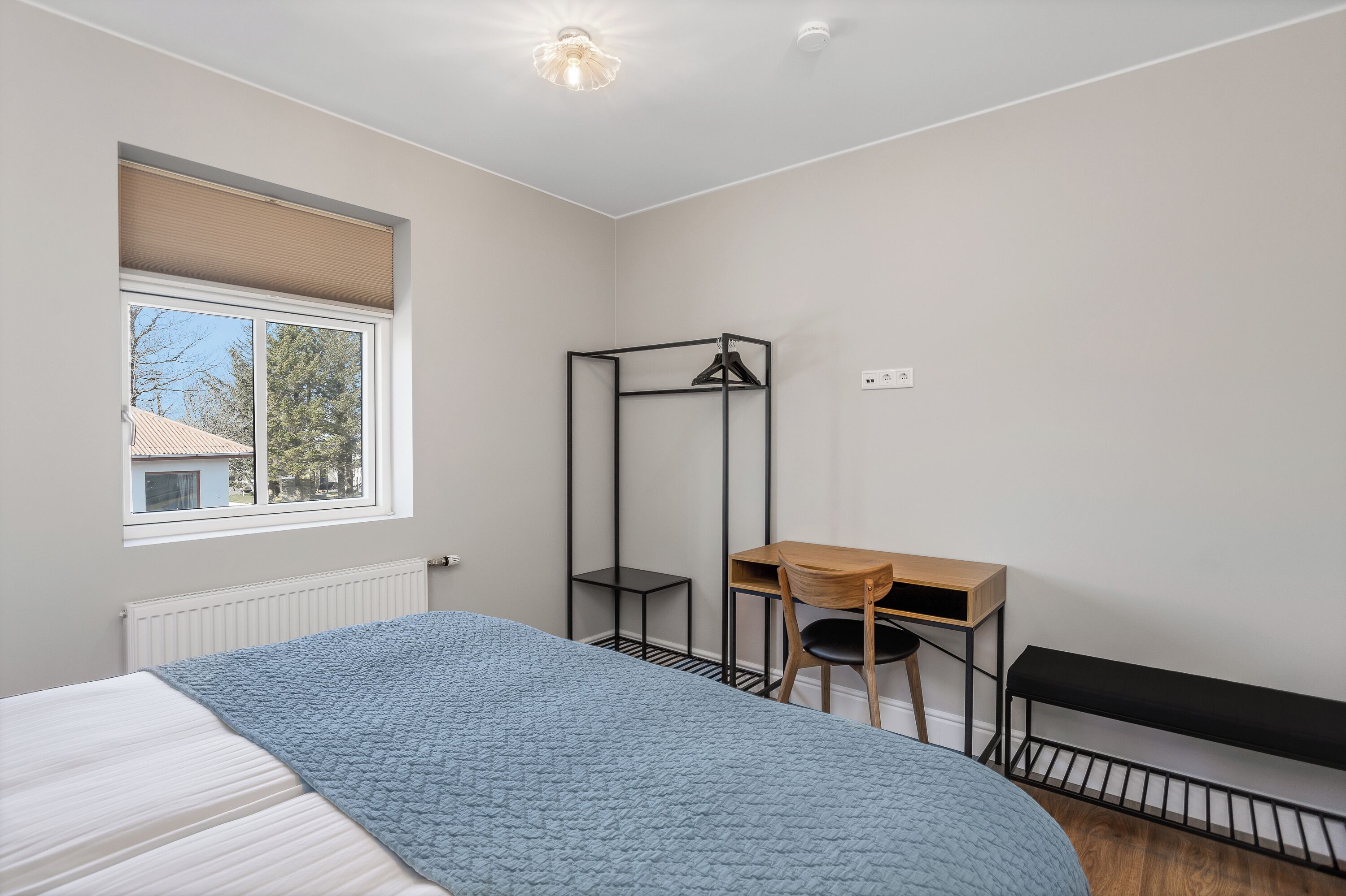 Une chambre à coucher comprenant un lit, un bureau, une chaise, un radiateur, une fenêtre avec des stores et un luminaire suspendu.
