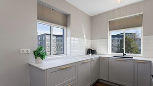 A kitchen with grey cabinets, a white countertop, a plant, and a window with blinds. - Aska Selfoss (Selfoss)