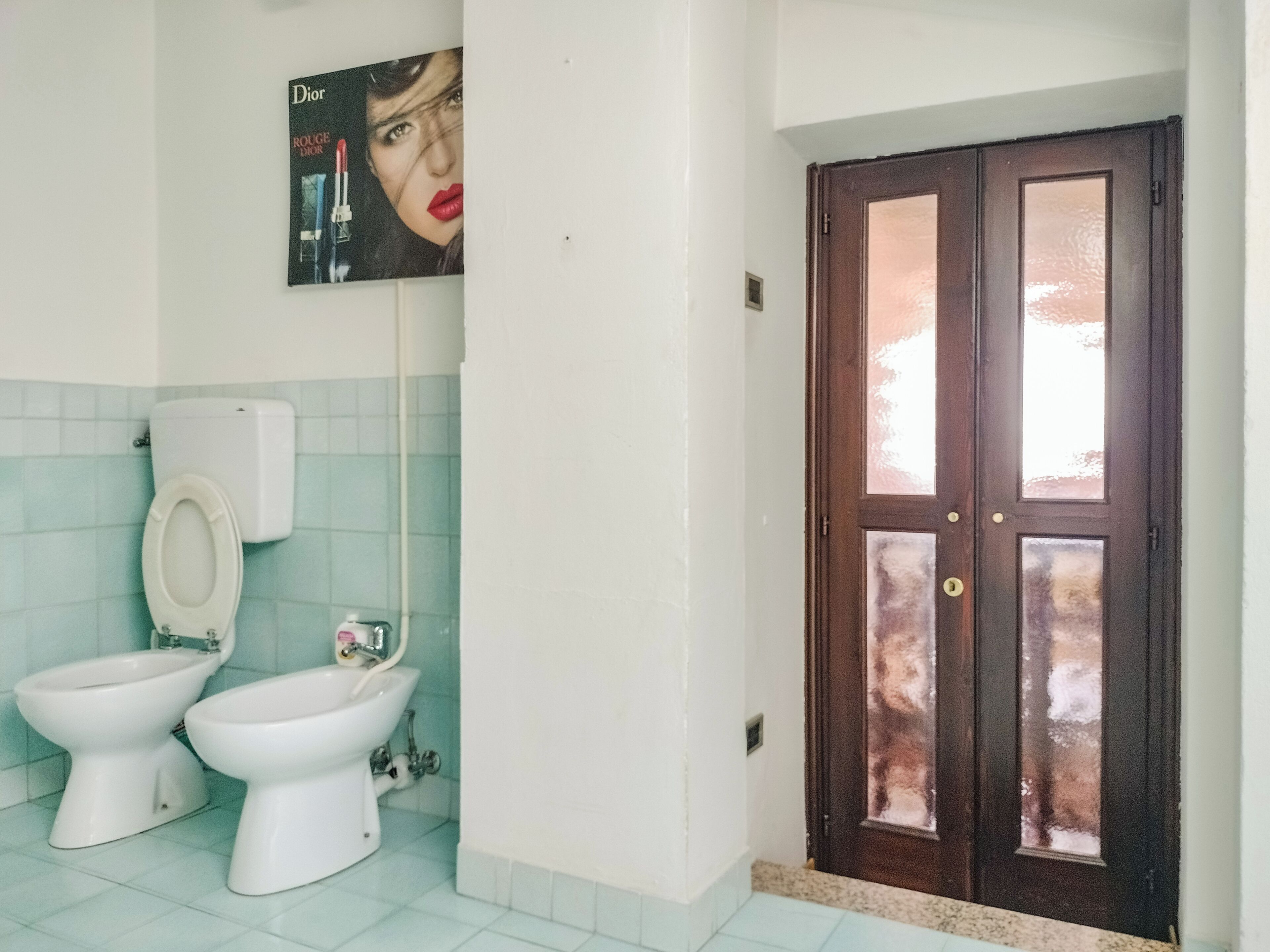 A bathroom with two toilets, a bidet, a toilet paper holder, and a door with glass panels.