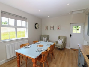 A dining area with a table set for four, wooden chairs, a radiator, a clock, and framed pictures on the wall. - Keepers Cottage (Haddington)
