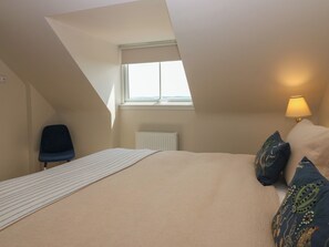 A bedroom with a sloped ceiling, a window, a radiator, a bed with a striped bedspread, a chair, and a lamp.
