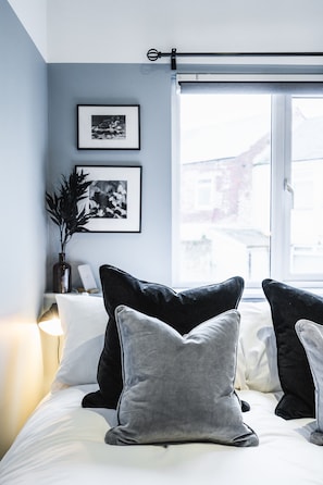 Ein Schlafzimmer mit Bett, Kissen, Nachttisch, Lampe, Fenster und zwei gerahmten Bildern an der Wand.