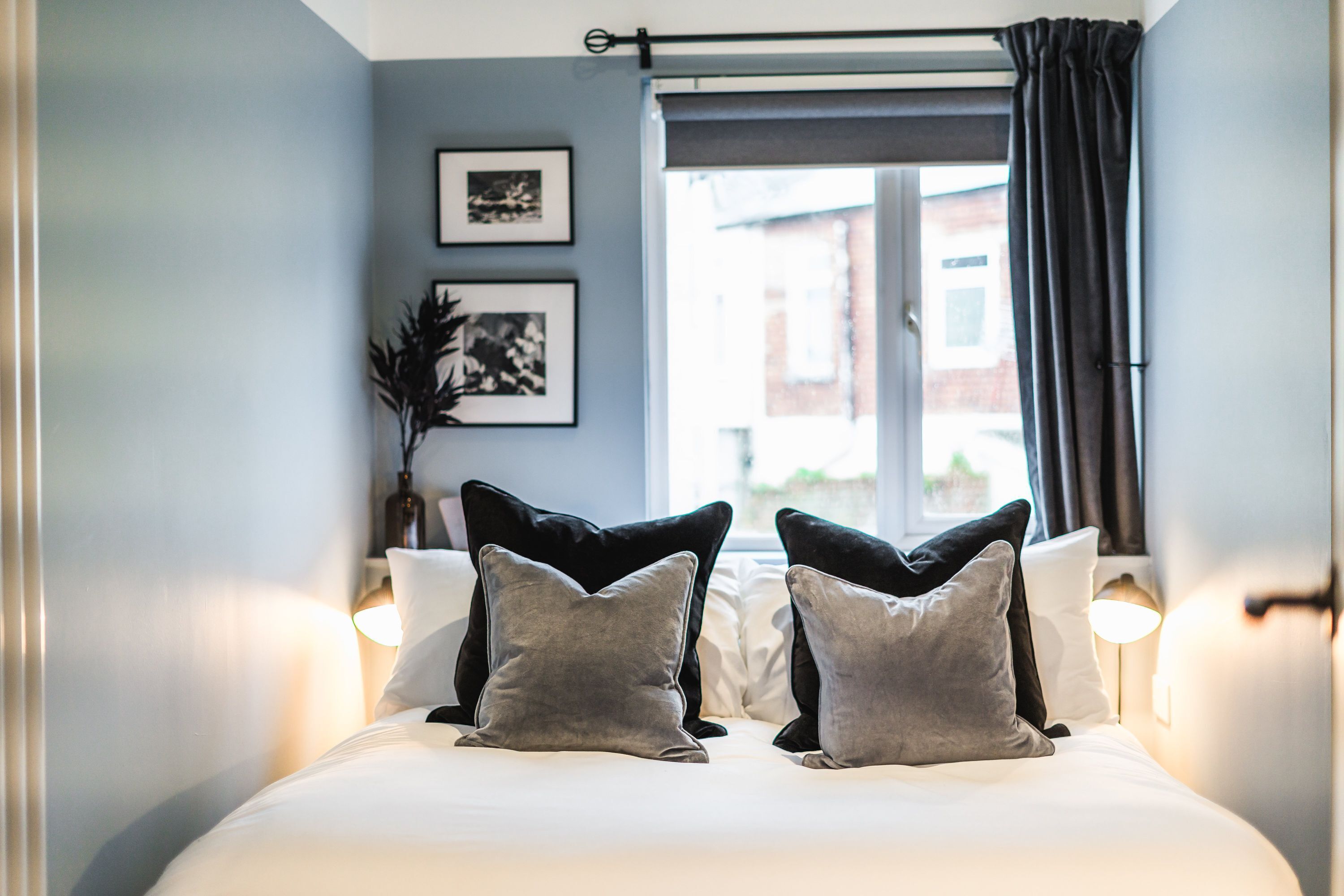 A neatly made bed with pillows, a window with curtains, and framed pictures on the wall.