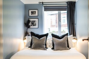 Ein ordentlich bezogenes Bett mit Kissen, ein Fenster mit Vorhängen und gerahmte Bilder an der Wand.