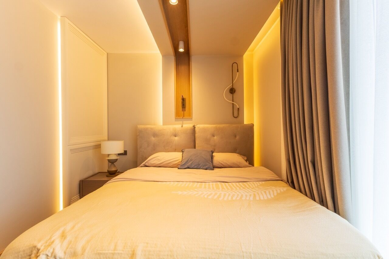 A hotel room with a large bed, a nightstand with a lamp, a wall-mounted light fixture, and a window with curtains.