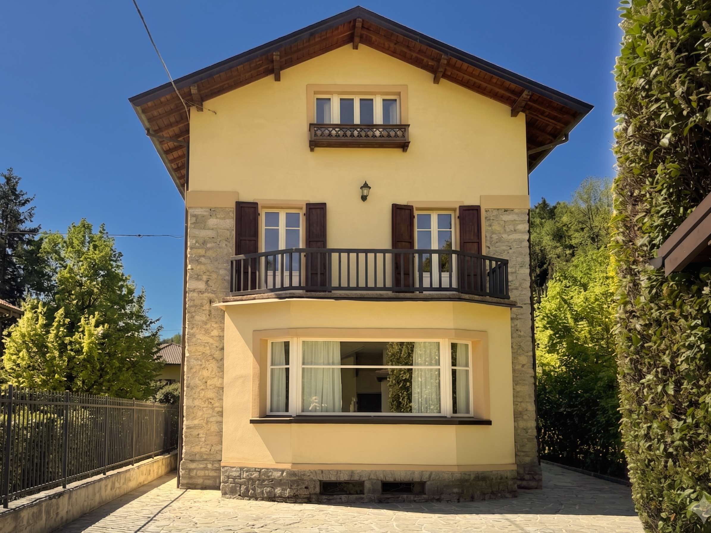 A two-story building with a balcony, stone base, and a window with curtains.