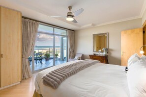 Habitación de hotel con una cama grande, ventilador de techo, vista a la playa y puerta corrediza de vidrio.