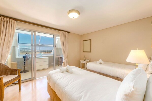 Habitación de hotel con dos camas, una zona de estar con una silla, una mesita y un ventanal con cortinas.