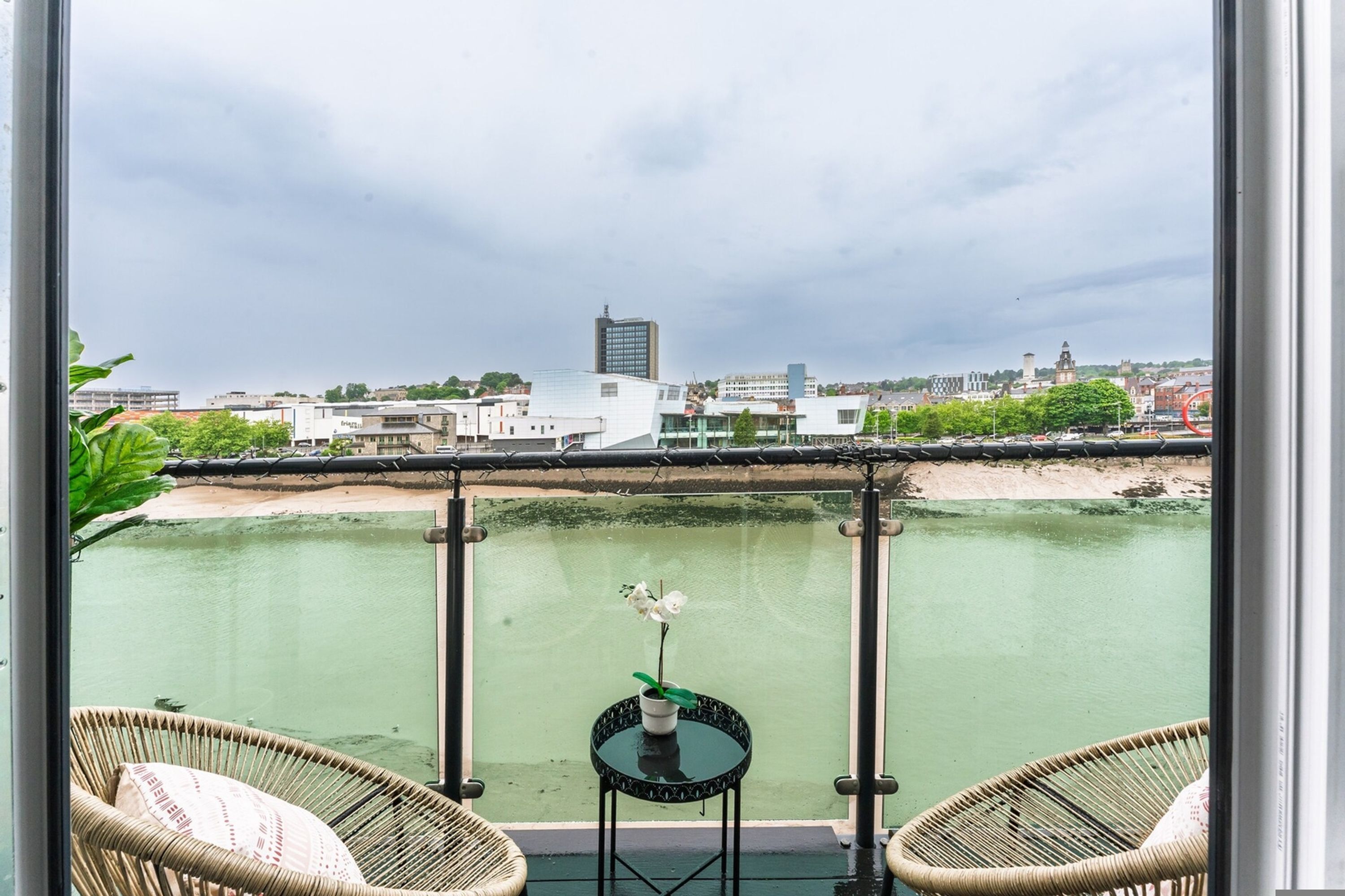 Ein Balkon mit Korbstühlen, einem kleinen Tisch mit einer Vase und Blick auf Stadt und Fluss.