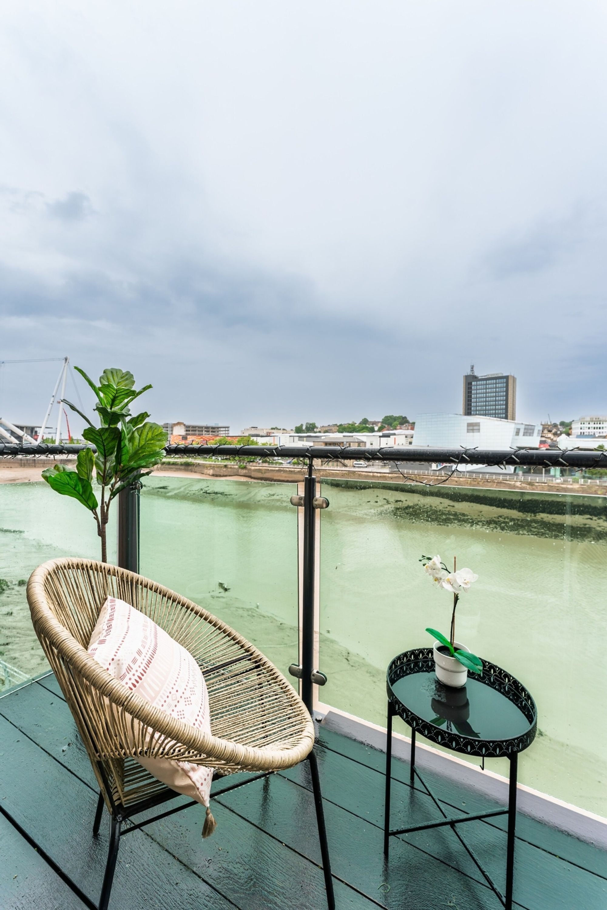 A balcony with a wicker chair, a small table with a potted plant, and a view of a river and buildings.
