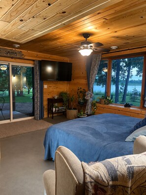Ein Zimmer in einer Holzhütte mit einem Bett, einem Fernseher, einem Schreibtisch und Blick ins Freie.