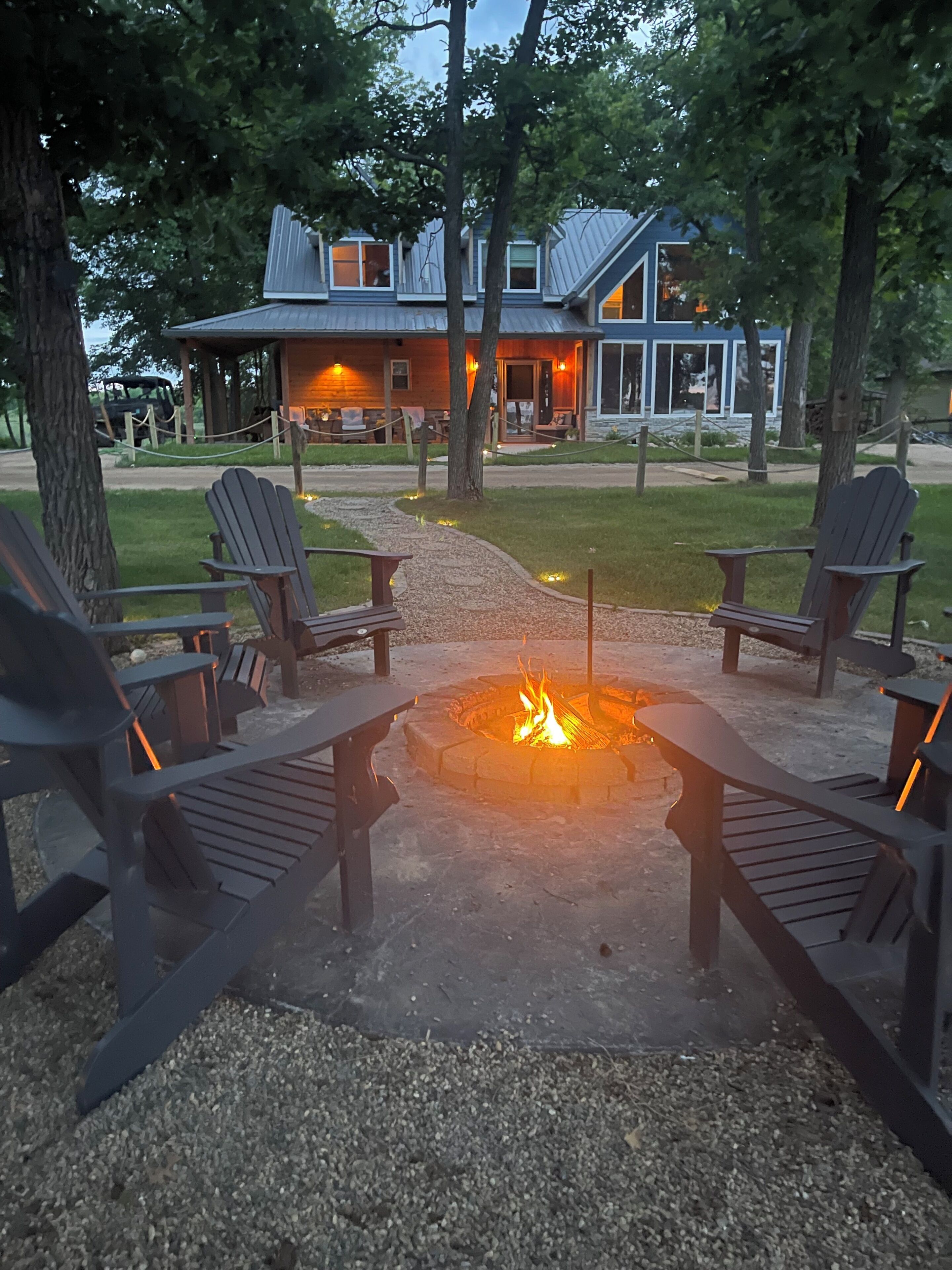 A fire pit with Adirondack chairs, a building with a metal roof, and a gravel path.