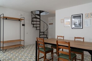 Un comedor con una escalera espiral, sillas de madera con cojines verdes y suelo de baldosas.