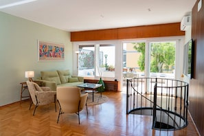 Un salón amplio con suelo de madera, un sofá verde, un sillón estampado, una mesita y una escalera de caracol.