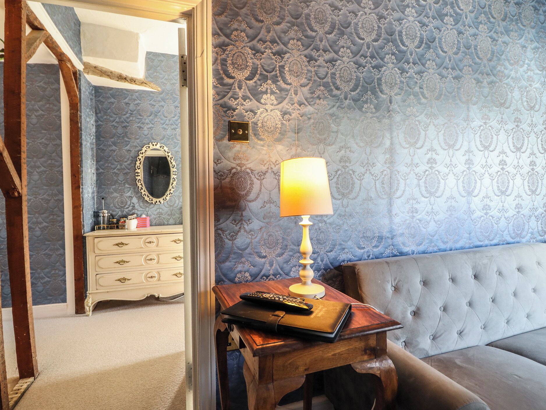 A room with a patterned wallpaper, a wooden dresser, a mirror, a lamp, and a tufted sofa.