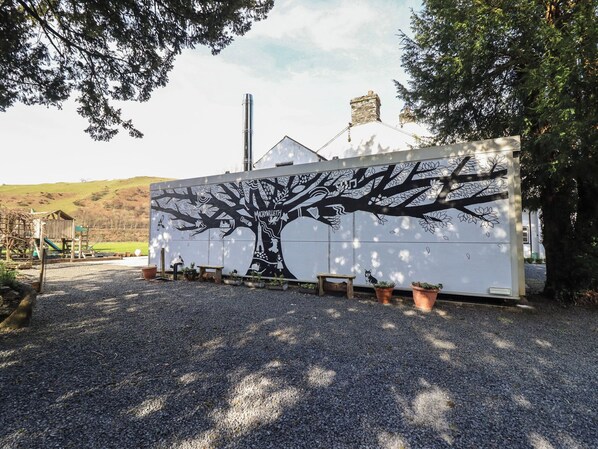 A building with a large tree mural on its side, gravel ground, and a wooden play structure in the background.