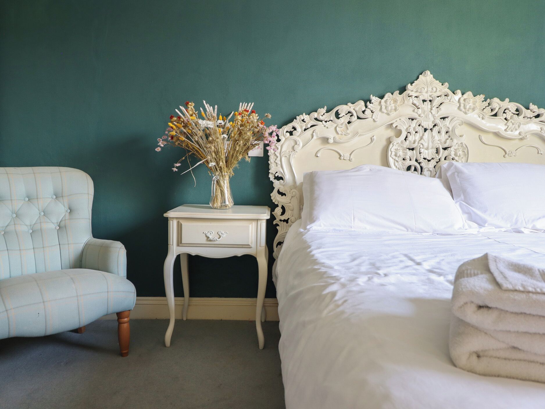 Una camera da letto con un letto, un comodino, una sedia e un vaso di fiori secchi.