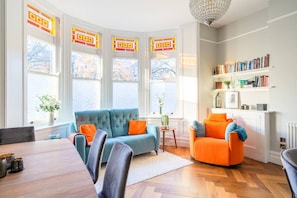 Ein modernes Wohnzimmer mit einem blauen Sofa, einem orangefarbenen Sessel, einem Esstisch und einem Bücherregal.