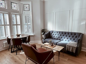 Ein Wohnzimmer mit grauem Sofa, Couchtisch, bequemen roten Sessel, Esstisch mit Stühlen und einem großen Fenster mit Rollläden.