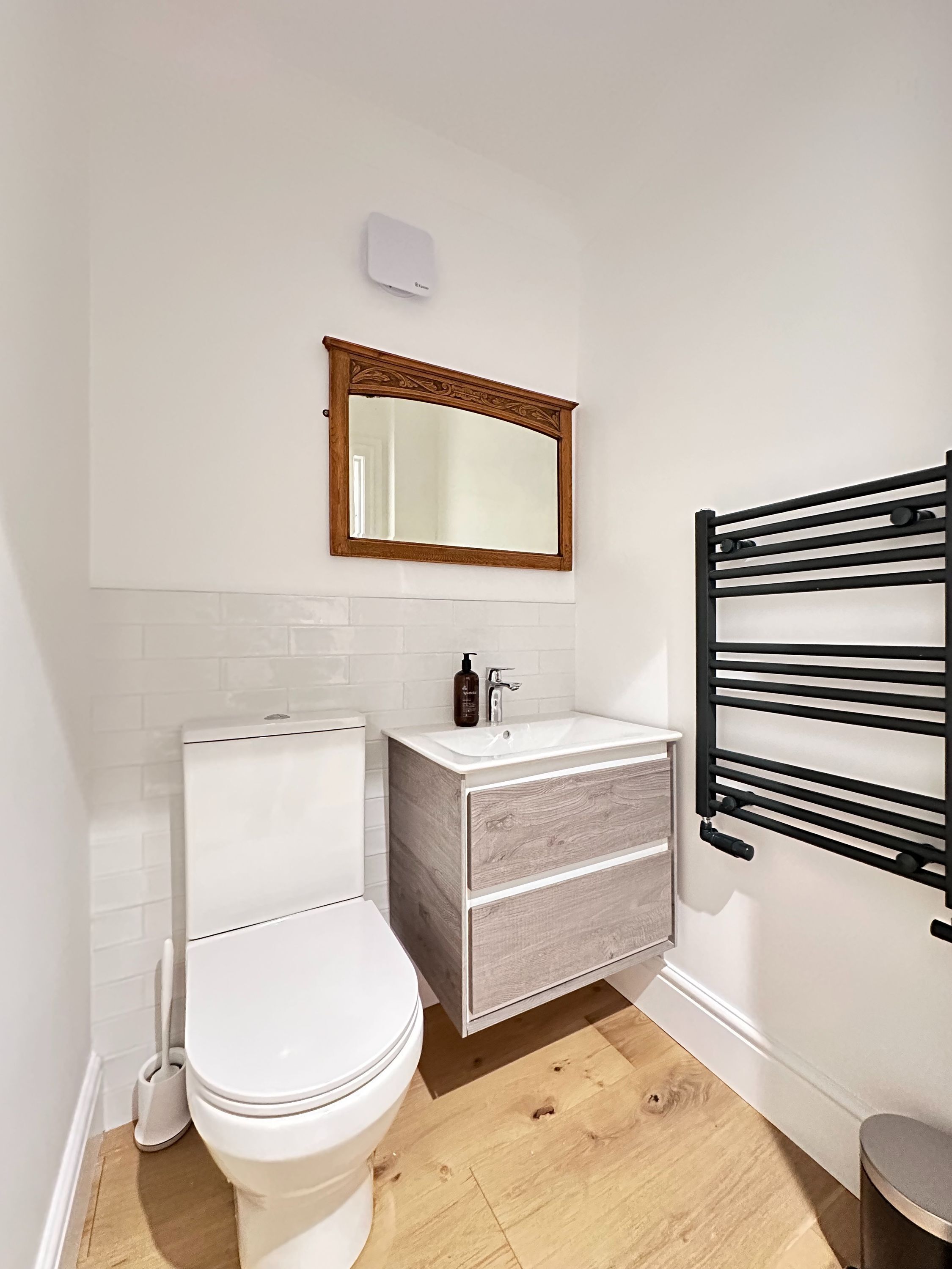 Une salle de bain avec des toilettes blanches, un meuble-lavabo en bois avec un miroir et un porte-serviettes noir.