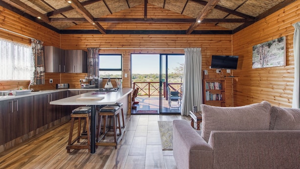 A wooden cabin interior with a kitchen, dining area, and a living room with a sofa.